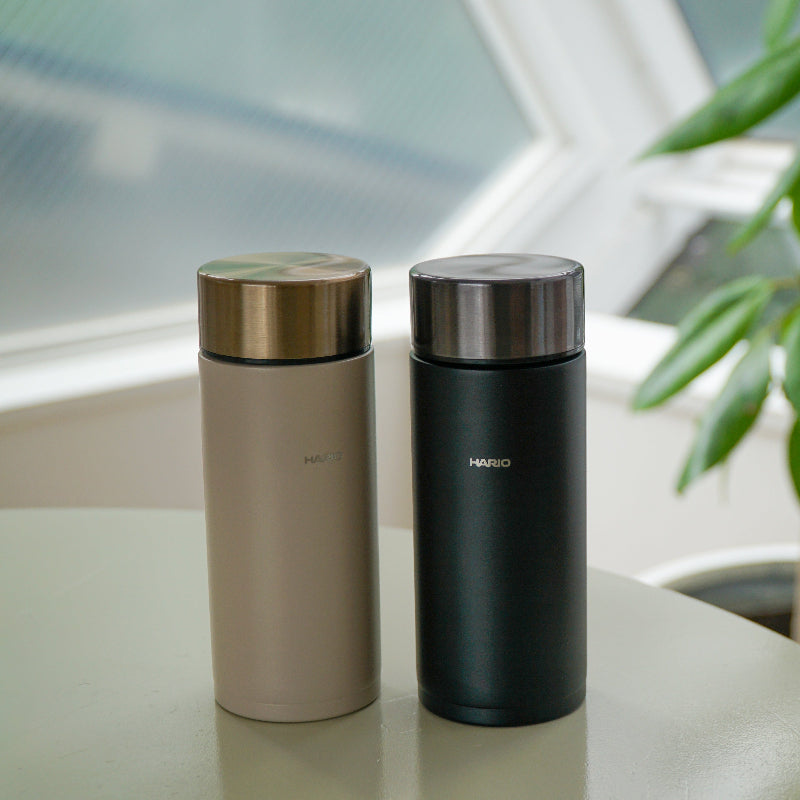 Two HARIO brand vacuum insulated tumblers standing on a light-coloured table near a window, one in rose gold and one in matte black, with a blurred green plant in the background.